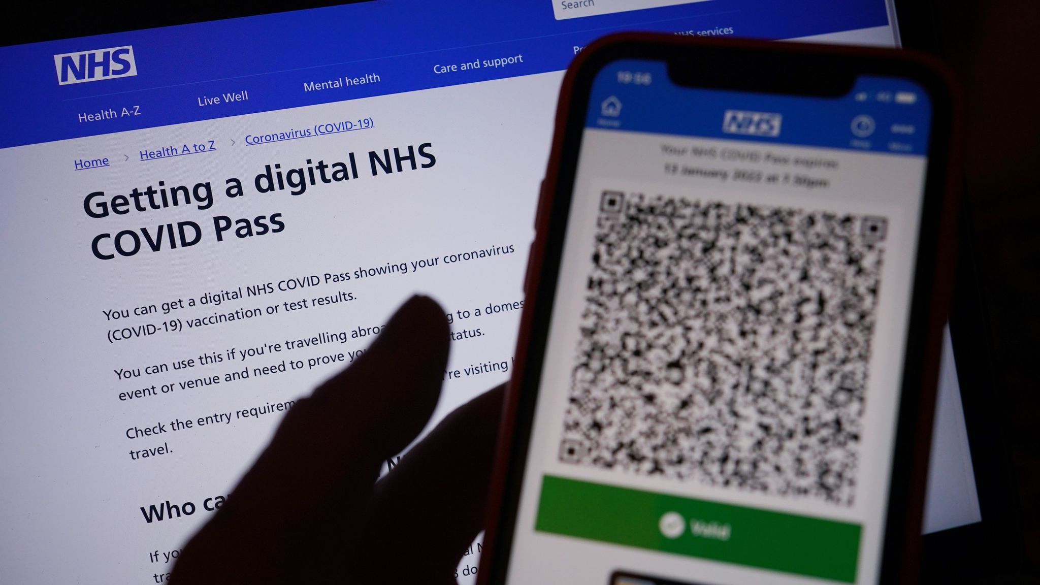 A hand holds a mobile phone shoes an NHS covid pass, with the NHDS webpage about how to get a digital covid pass in the background
